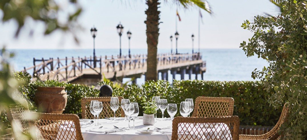 Tables laid on a terrace by the sea, with a wooden pier beyond