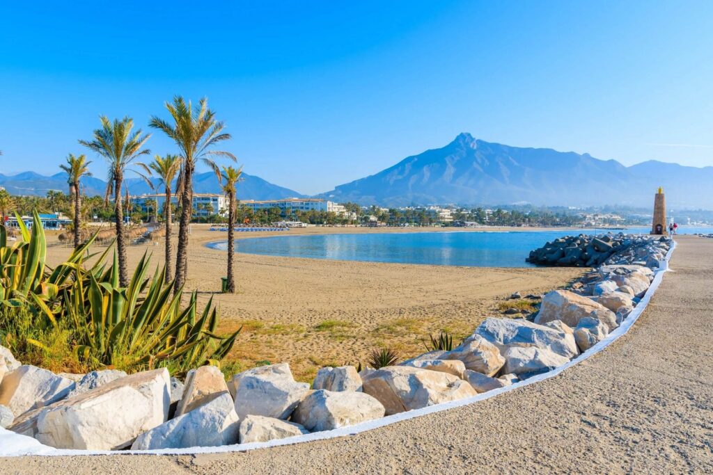 A sandy Marbella beach with palms, rocks and La Concha in the distance